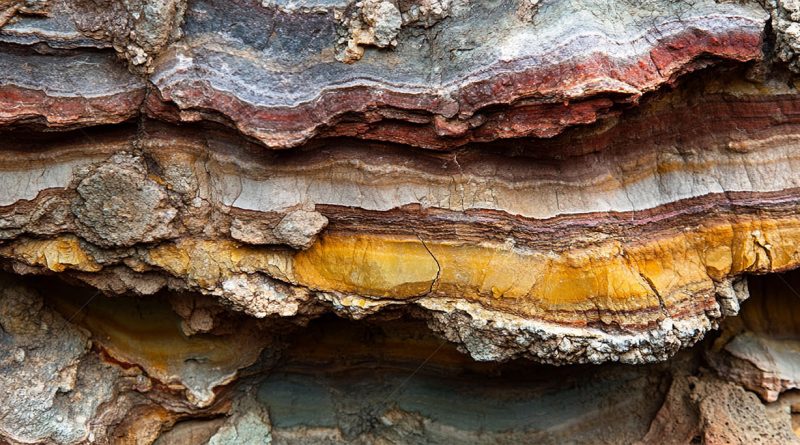 Close-up of layered mineral-rich rock formation showing distinct bands of red, yellow, and grey mineral deposits representing common minerals in Earth’s crust.