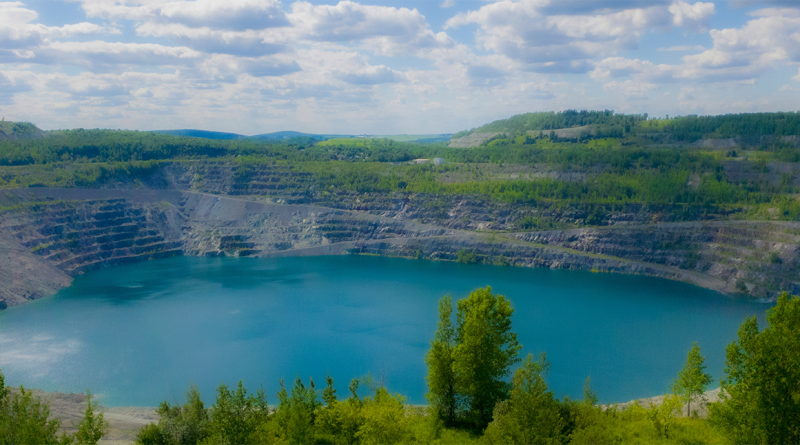 Wide view of a terraced open-pit mine in Quebec filled with turquoise water, surrounded by forested hills under a cloudy sky.