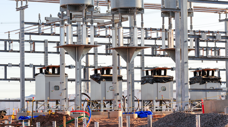 A construction site for a battery metal project featuring industrial steel scaffolding, electrical control boxes, and heavy infrastructure under bright sunlight.